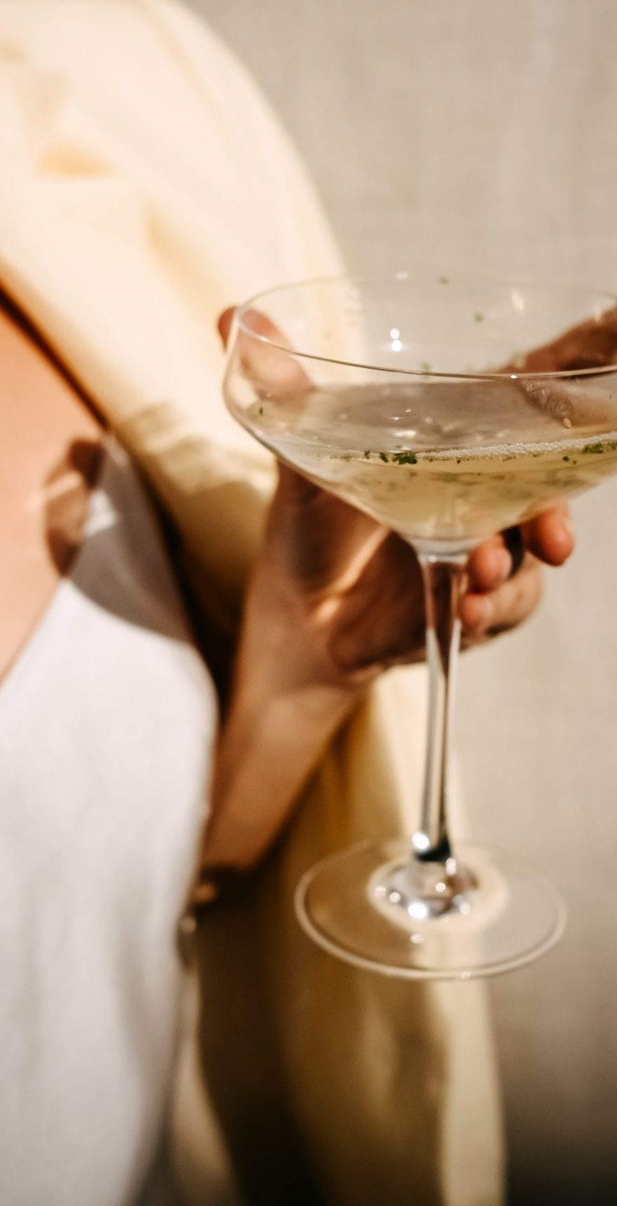 Woman dressed in white dress holding a coupe with a cocktail in sun light. Concept of an open air party.